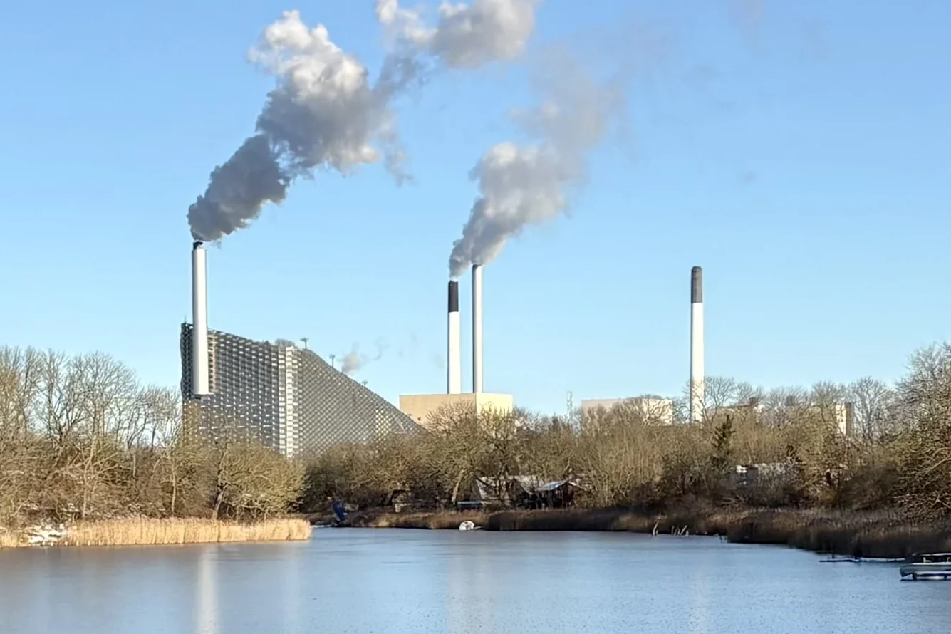 An industrial facility with chimneys, trees and water in the foreground.